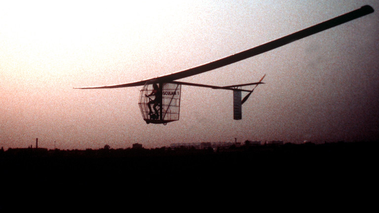 Ein Mann sitzt in einem Käfig auf einer Art Fahrrad, an das 20 Meter breite Flügel und ein Heck mit Propeller montiert sind
