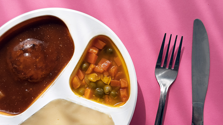 Foto einer Fertigmahlzeit mit Frikadelle, Gemüse und Kartoffelpüree in einem Plastikteller, daneben liegt Besteck