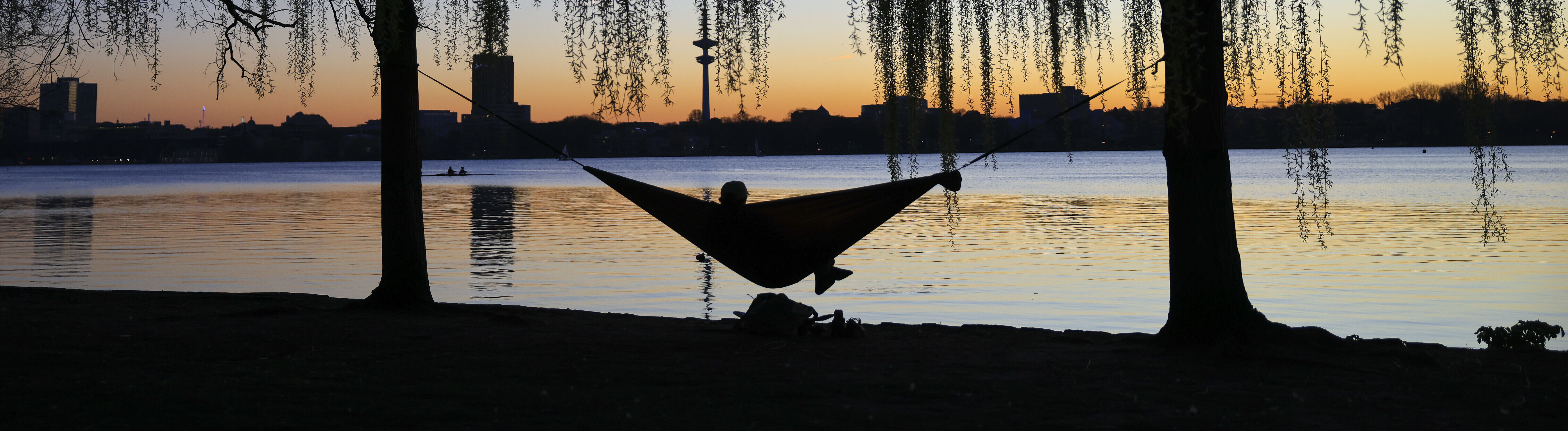 Ein Mensch liegt an einem See in einer Hängematte. Im Hintergrund sind Gebäude erkennbar. Die Sonne geht gerade unter.