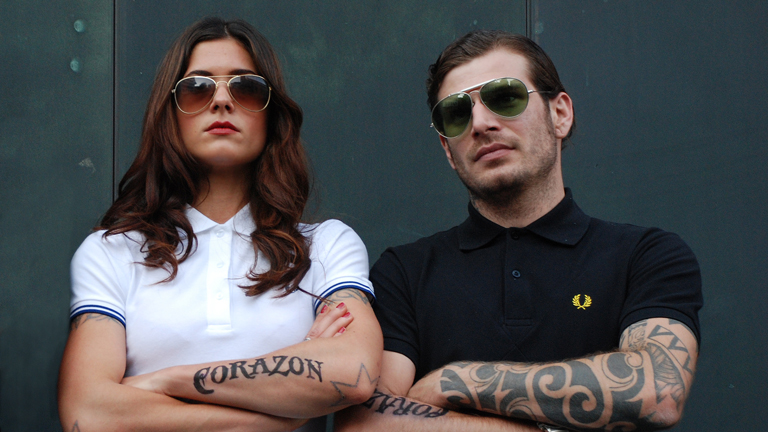 Eine Frau und ein Mann stehen provokant mit verschränkten, tätowierten Armen da und tragen Sonnenbrillen.