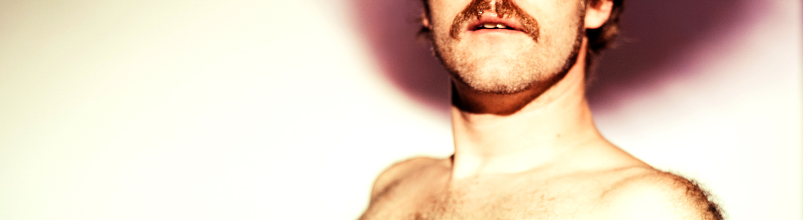 Ein behaarter Mann mit Schnauzbart trägt ein enges Oberteil. Man sieht seine Brust.