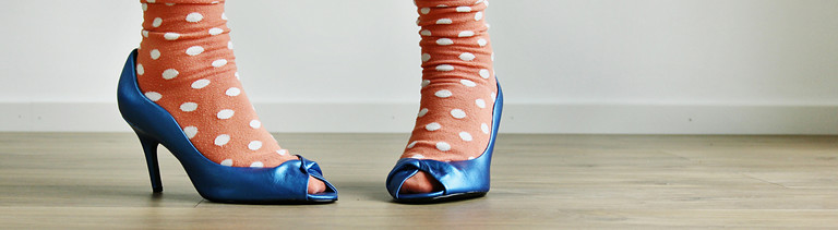 Zwei Füße in blauen Pumps mit orangenen Socken mit weißen Punkten darauf.