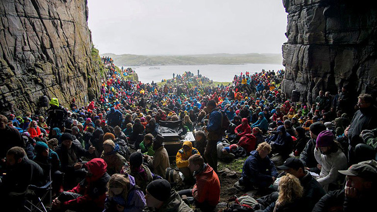 In einer großen Höhle sitzen viele Menschen mit Regenkleidung bekleidet mit Blick auf das Wasser.