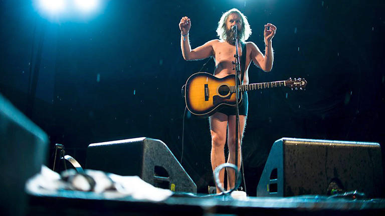 Ein Mann in Unterhose steht auf der Bühne. Er hat lange Haare und Bart und eine Gitarre umhängen.