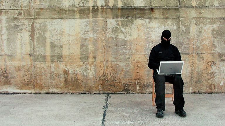 Symbolbild eines Hackers der vermummt mit LapTop auf einem Stuhl sitzt.