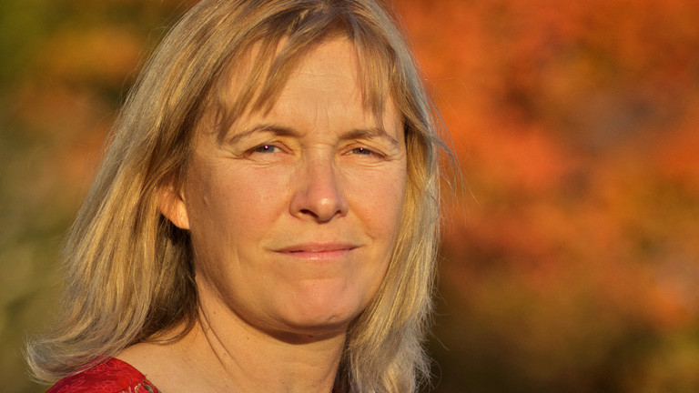 Portrait von Valerie Curtis. Sie hat schulterlange, blonde Haare. Herbstsonne und gefärbte Bäume im Hintergrund.