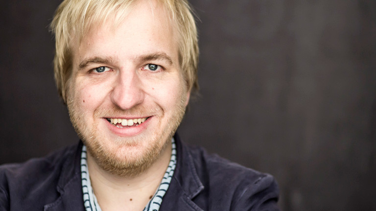 Portrait von David Denk. Er hat blonde kurze Haare und trägt Bart.
