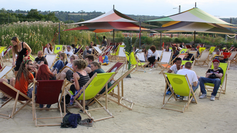 Auf einem Platz mit Sand sitzen viele junge Leute in Strandliegen. Ein paar große Sonnenschirme stehen zwischen den Stühlen.