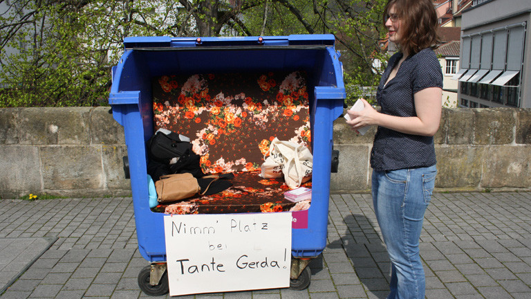 Eine blaue Papiermülltonne steht auf einem städtischen Platz. Die Tonne ist vorne aufgeschnitten. In das Innere ist eine bunte Sitzbank gebaut.