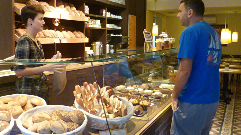 Blick in den Verkaufsraum von Annas Bakery. In der Auslage liegen Brötchen, Laugenwerk. Anna bedient einen Kunden.