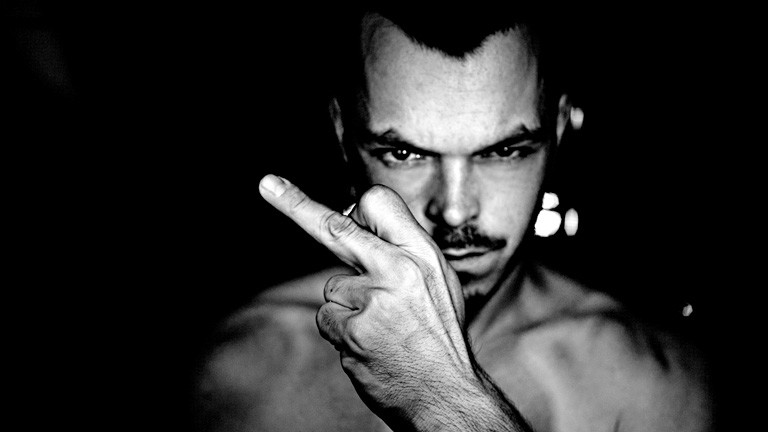 Schwarz-Weiß-Foto. Ein Mann blickt finster in die Kamera, mit nacktem Oberkörper. Er hält seine linke Hand vor sich mit ausgestrecktem Mittelfinger. Der Hintergrund ist schwarz.