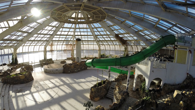 Blick in eine leerstehende Schwimmhalle mit Glaskuppel und verglasten Außenseiten. Unter der Kuppel war einmal das Wellenbad. Rechts ist eine grüne Rutschbahn.