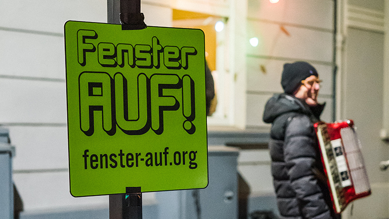 Auf einem Schild steht "Fenster auf!", dahinter ein Ziehharmonika-Spieler.