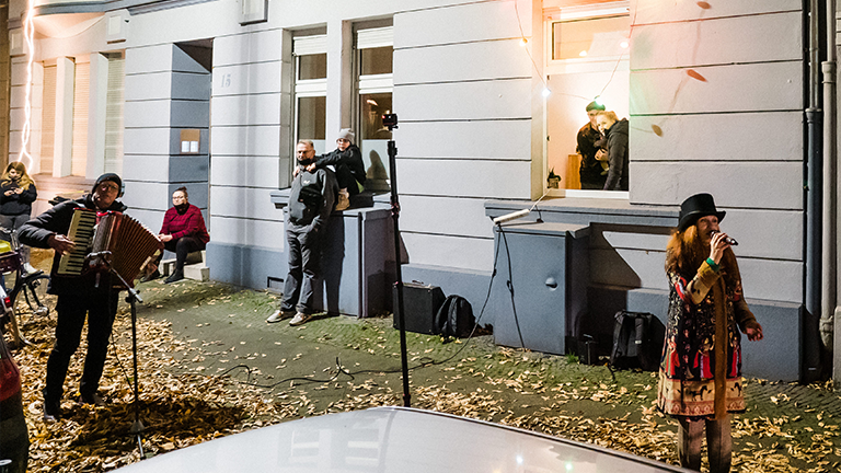Eine Band spielt auf einem Bürgersteig, dahinter neugierige Menschen, die aus dem Fenster schauen.