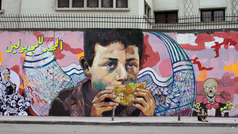 A young boy (homeless) in tears holding a sandwich who is Martyr Sayed Khaled the youngest Martyr in the Revolution, Arabic says “glory to the unidentified”