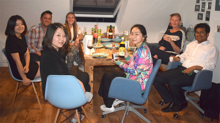 An einem Tisch sitzen acht Männer und Frauen. Auf dem Tisch stehen Teller, Gläser und Essen.