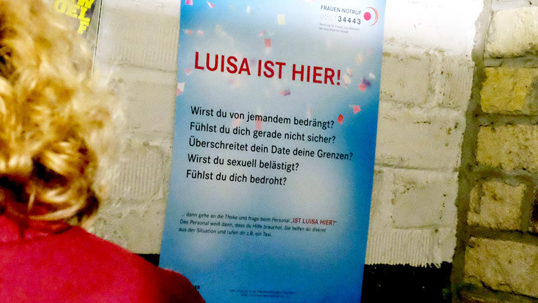In einer Kneipe hängt ein Plakat der Kampagne "Luisa hier"