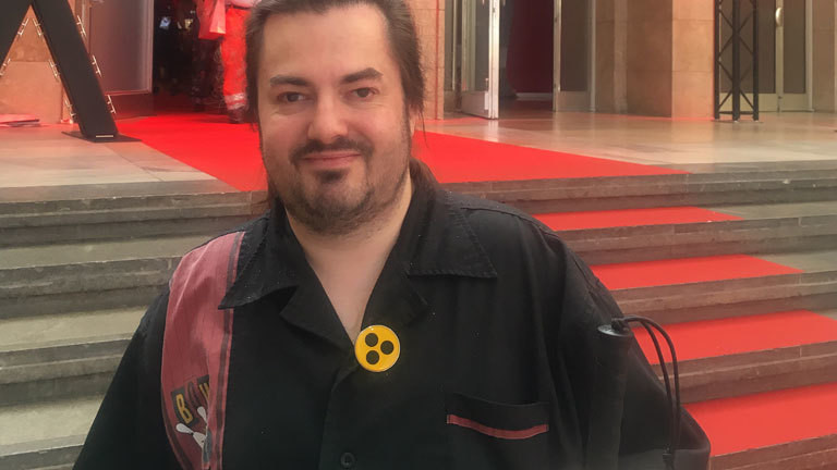 Tim ist 37 Jahre alt, trägt einen langen braunen Zopf und ein schwarzes Hemd. Auf der Vorderseite trägt Tim ein kleines rundes Blindenabzeichen.