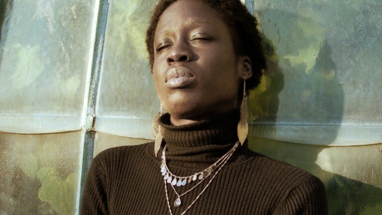 Eine Frau lehnt an einer Wand, hat ihre Augen geschlossen und entspannt.