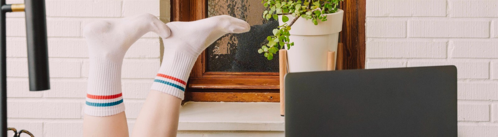 Gelangweilte Frau mit hochgelegten Füßen am Laptop am Tisch im Homeoffice.