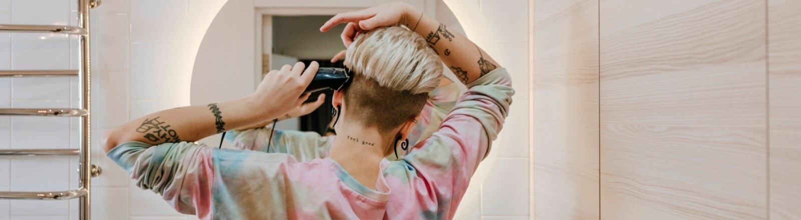 Eine Frau steht in einem Badezimmer vor einem Spiegel. Wir sehen sie von hinten. Sie setzt am Kopf eine Haarschneidemaschine an.