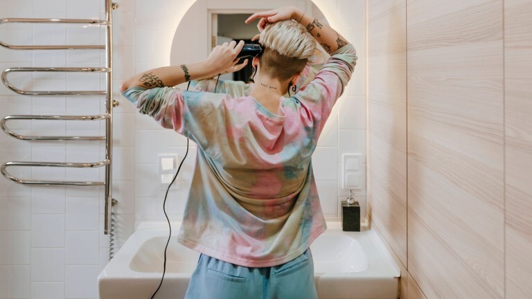 Eine Frau steht in einem Badezimmer vor einem Spiegel. Wir sehen sie von hinten. Sie setzt am Kopf eine Haarschneidemaschine an.