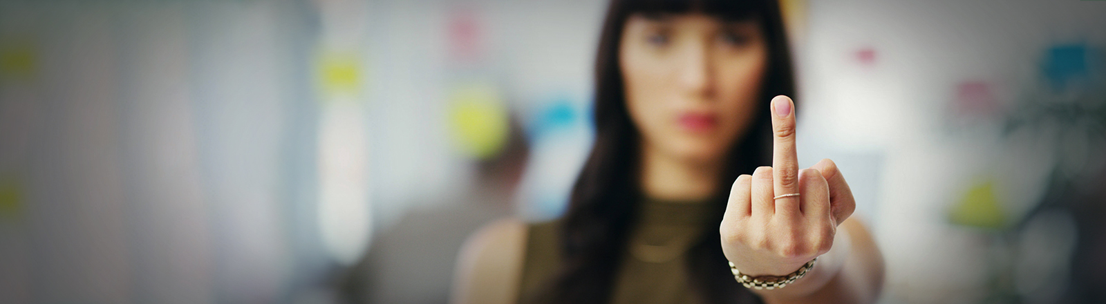 Foto einer jungen Frau in einem Büro, die eine Hand mit einem ausgestrecktem Stinkefinger in die Kamera hält und selbst im Hintergrund unscharf ist