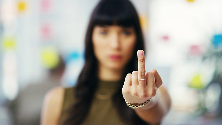 Foto einer jungen Frau in einem Büro, die eine Hand mit einem ausgestrecktem Stinkefinger in die Kamera hält und selbst im Hintergrund unscharf ist