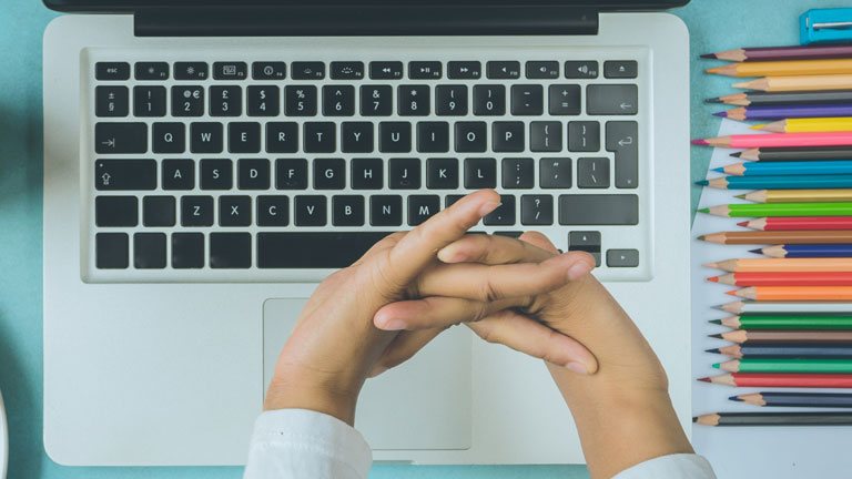 Hände über einer Laptop-Tastatur, daneben Buntstifte