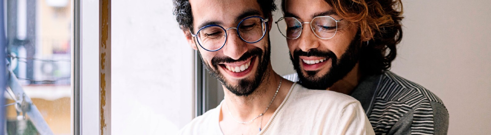 Ein Mann umarmt einen anderen Mann von hinten. Sie schauen beide gemeinsam auf ein Handy und lächeln dabei.