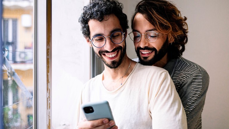 Ein Mann umarmt einen anderen Mann von hinten. Sie schauen beide gemeinsam auf ein Handy und lächeln dabei.