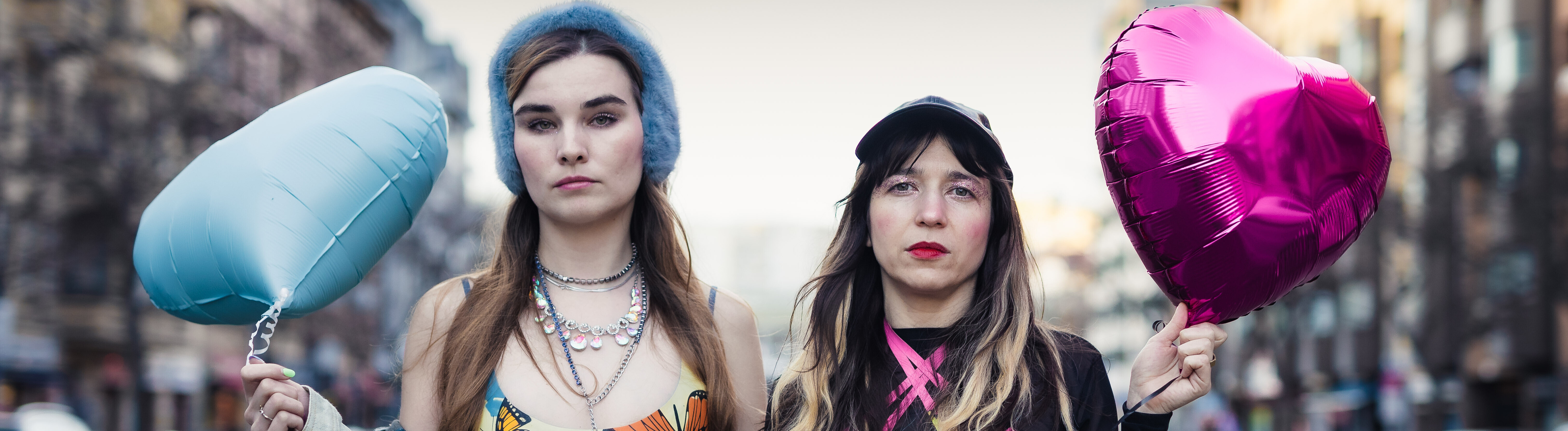 Pauline Schmiechen und Paulita Pappel stehen zwischen zwei Straßen in Berlin, sie tragen bunte Kleidung und einen blauen und einen pinken herzförmigen Luftballon