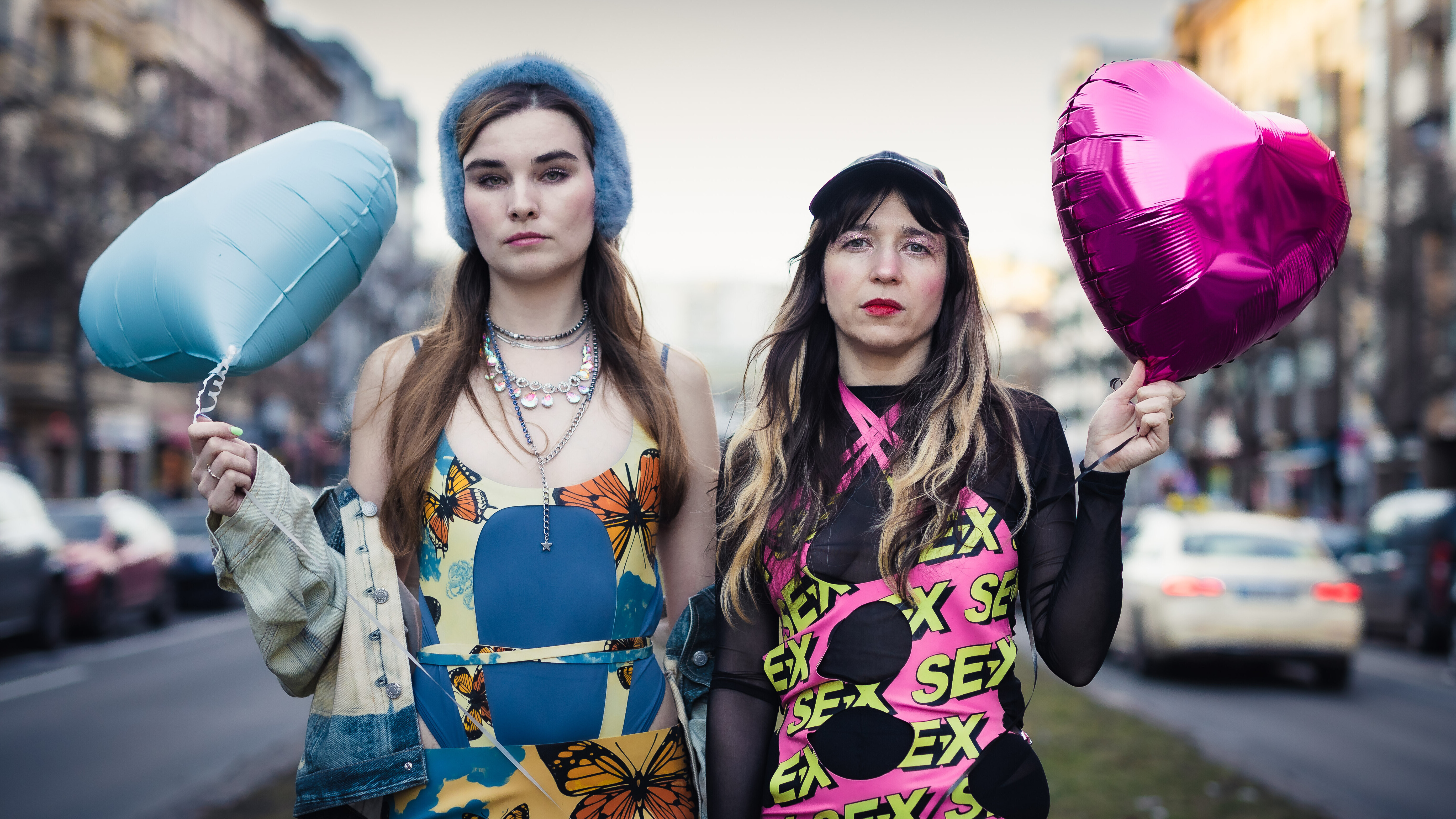 Pauline Schmiechen und Paulita Pappel stehen zwischen zwei Straßen in Berlin, sie tragen bunte Kleidung und einen blauen und einen pinken herzförmigen Luftballon