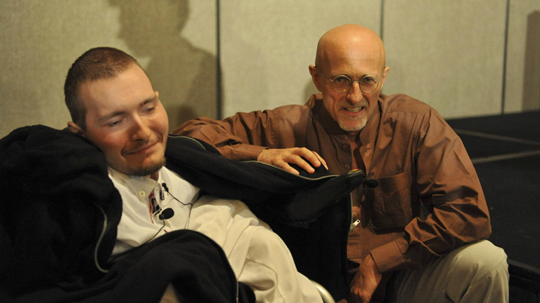 Waleri Spiridonow sitzt in einem Rollstuhl, er trägt ein weißes Hemd. Die Haare sind kurz. Neben ihm kniet der Chirurg Sergio Canavero.