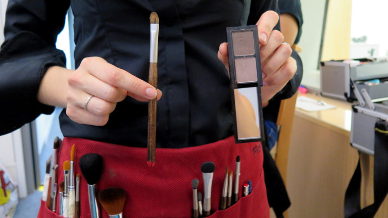 Florance hält Pinsel und zwei dunkle braune Lidschatten in die Kamera