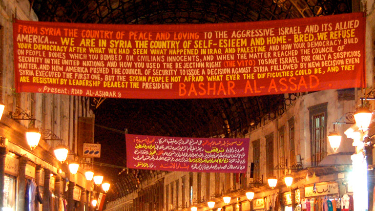 Auf einem Banner steht ein Text, Propaganda der syrischen Regierung. Er beginnt mit: Aus Syrien, dem Land des Frieden und der Liebe.