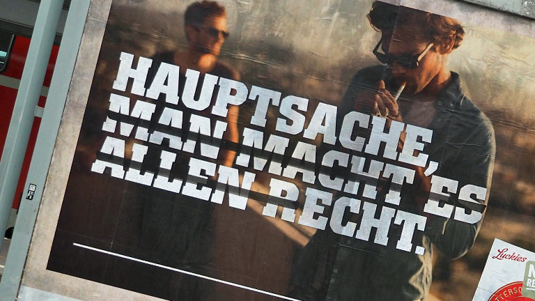 Auf einem Bahnhof hängt ein Werbeplakat für Zigaretten: "Hauptsache (macht macht es allen r)echt".