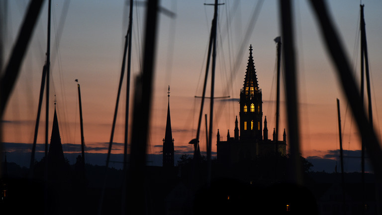 Der Turm des Münsters in Konstanz (Baden-Württemberg) wird am 28.09.2015 kurz nach Sonnenuntergang von Innen beleuchtet. Im Vordergrund sind die Masten mehrerer Segelboote zu sehen, die im Yachthafen von Konstanz liegen.