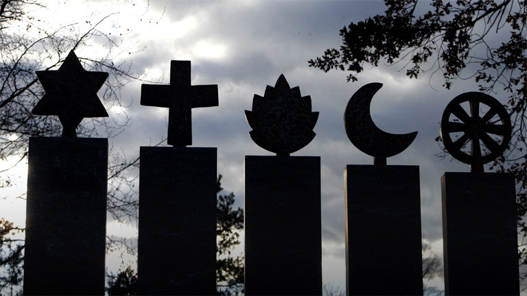 Die Symbole der Religionen Judentum, Christentum, Hinduismus, Islam und Buddhismus aus Stein gehauen nebeneinander.