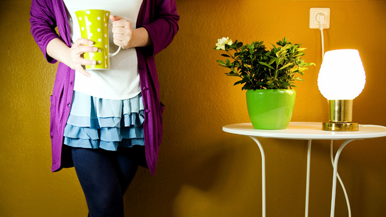 Ein Frauenkörper mit Kaffee in der Hand, auf einem Tisch eine Blume und eine leuchtende Lampe.