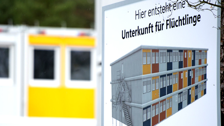Auf einem Schild steht eine Füchtlingsunterkunft als Containerbau abgebildet.