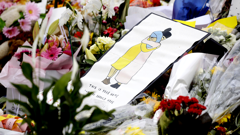 Zwischen Blumen liegt eine Zeichnung auf der sich zwei Menschen umarmen, darunter steht geschrieben: Das ist euer Zuhause, ihr hättet hier sicher sein sollen.