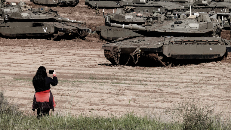Eine Frau mit einem Smartphone in der Hand steht vor jeder Menge Panzer auf einem Acker