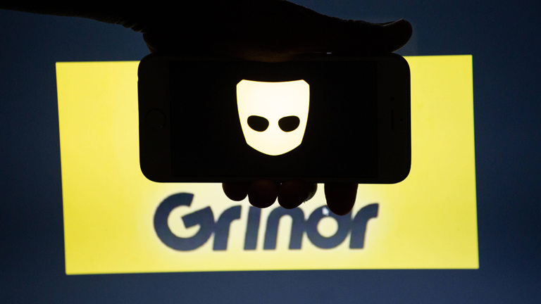Das Logo der App Grindr auf einem Smartphone, das von einer Hand vor einen Bildschirm gehalten wird auf dem der Schriftzu zu sehen ist.
