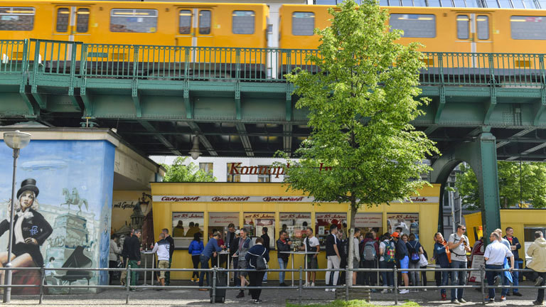 Blick auf "Konnopke's Imbiß" in Berlin an der Schönhauser Allee. Davor stehen Kunden Schlange. Der Imbiss ist unter der Hochbahn-Brücke einer U-Bahnlinie.