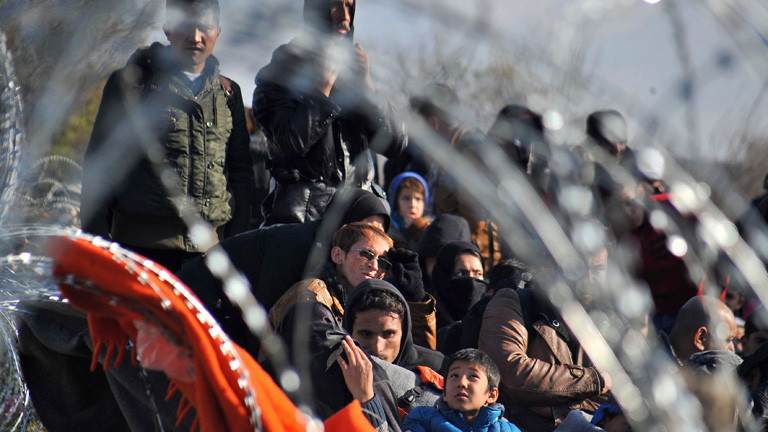 Eine Gruppe von Flüchtlingen an der griechisch-mazedonischen Grenze. Manche sitzen auf dem Boden, andere stehen. Vor ihnen ist Stacheldraht; Foto: dpa