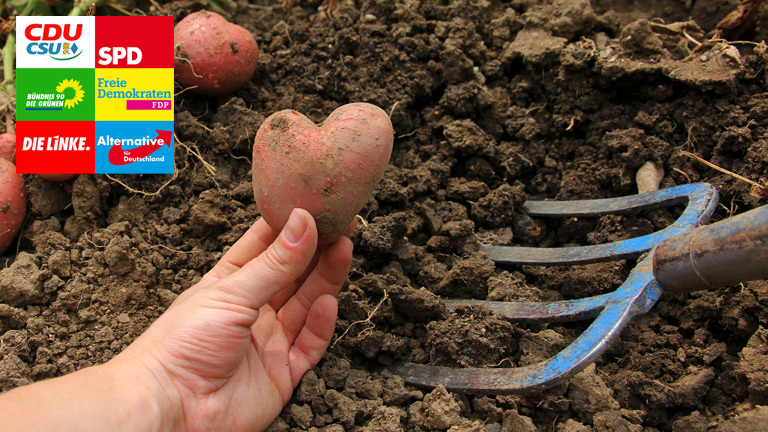Eine Hand hält eine herzförmige Kartoffel in der Hand. Dahinter liegt Erde, in der eine Harke steckt.