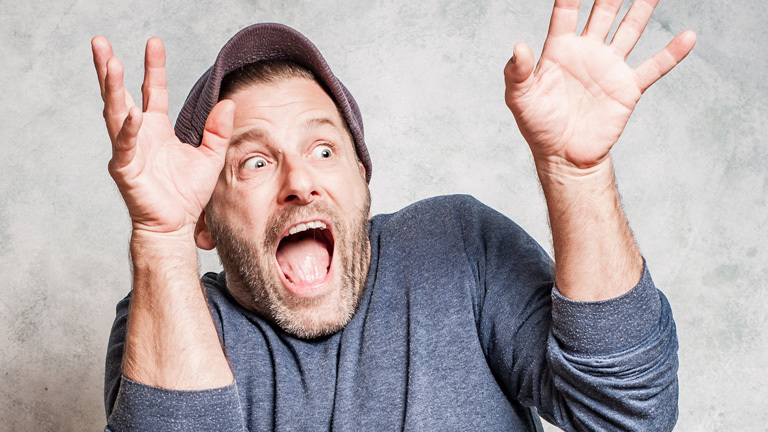 Ein Mann reißt die Hände nach oben und schaut erschrocken. Man sieht, dass er extra übertreibt.