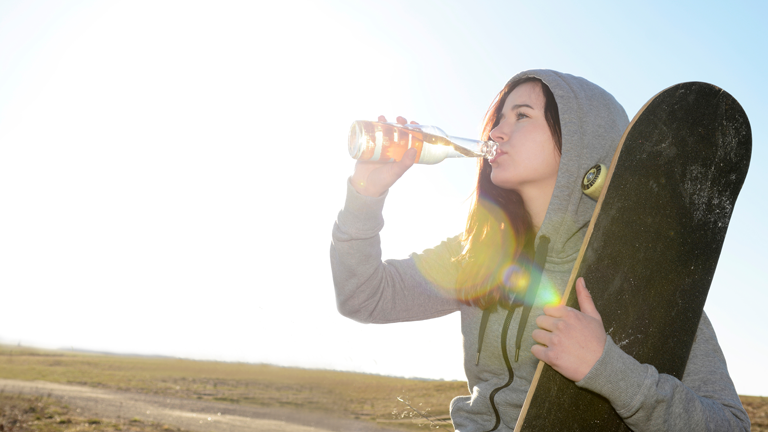 Ein Mädchen mit Skateboard unterm Arm trinkt aus einer Limoflasche