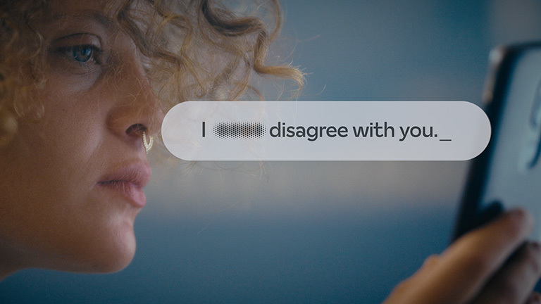 Eine Frau schaut auf ein Handy. Eingeblendet ist eine weiße Blase, in der steht "I disagree with you". Vor dem Wort "disagree" ist ein Wort verpixelt.
